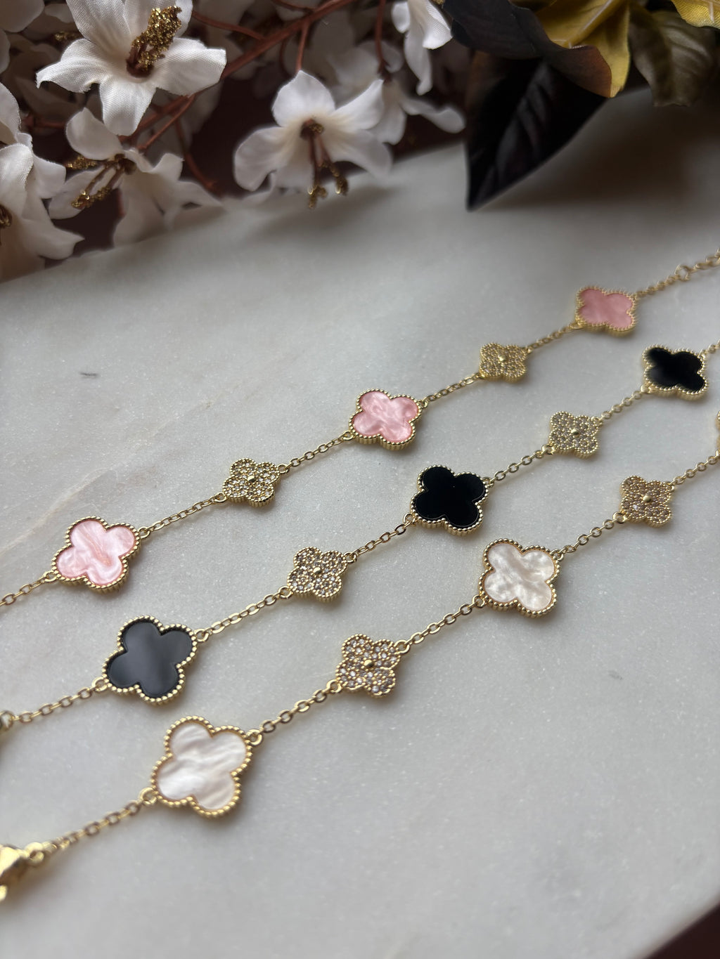 Drei vergoldete Kleeblatt-Armbänder mit weißen, roséfarbenen und schwarzen Anhängern aus Perlmutt auf rosa Samtkissen, umgeben von weißen Blüten.